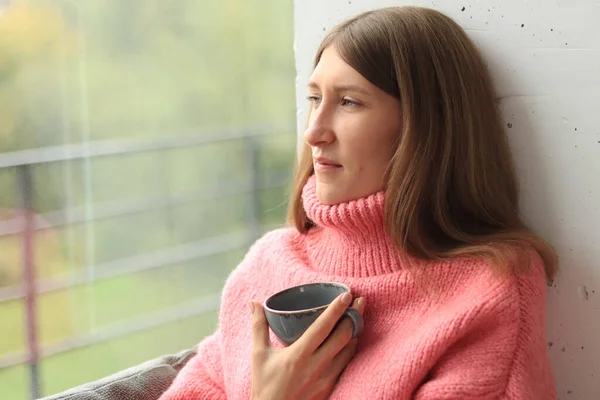 Rainy weather outside the window and a girl in a warm sweater looks at the rain. Girl with a cup in her hands, a cozy atmosphere of home comfort. Stay home. Autumn mood