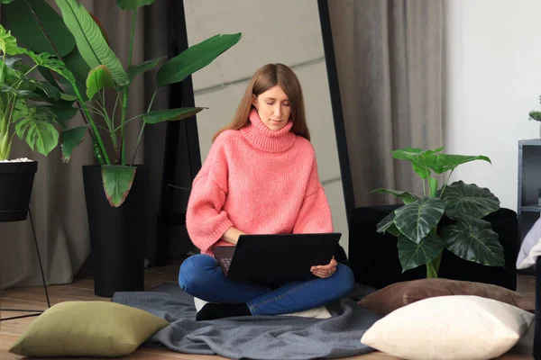 The girl works at a laptop at home in a cozy atmosphere. Remote work from home, blogging, communication via the Internet. Young woman working on a laptop while sitting on a pillow on the floor