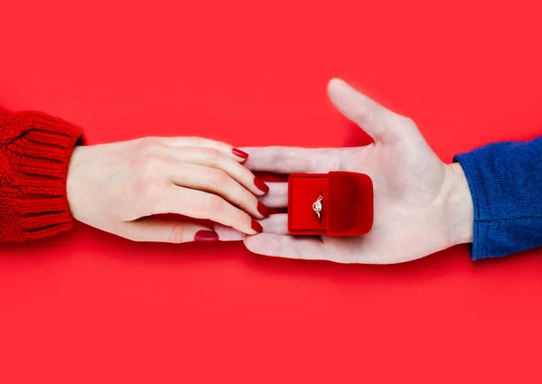 Man making marriage proposal. Male hand giving a gift box with engagement ring on red background. Close-up. Top view.