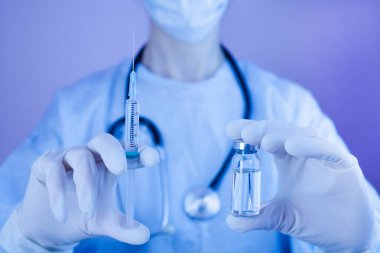Tıbbi maskeli ve koruyucu eldivenli kadın doktor ya da hemşire şırınga ve tıbbi enjeksiyonu ellerinde tutuyor. Grip ve koronavirüs aşısı. Neon renkli fotoğrafçılık. Yakın plan..