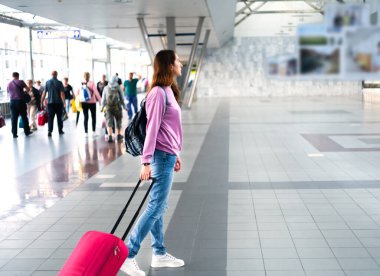 Sırt çantası ve kırmızı bavulu olan genç bir kadın havaalanında ya da tren istasyonunda duruyor. Boşluğu kopyala.