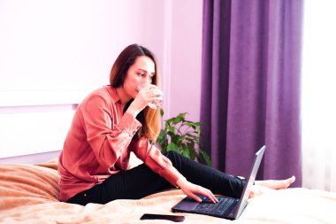 Genç bir kadın evdeki yatakta otururken su içer ve dizüstü bilgisayarla çalışır. Yakın plan. Seçici odak.