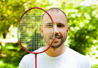 Genç ve çekici bir adam gülümsüyor ve güneşli bir günde parkta raket badmintonuyla yüzünü örtüyor. Sağlıklı yaşam tarzı kavramı. Yaşam tarzı, tatil, spor ve eğlence. Yakın plan..