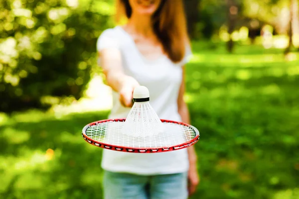 Beyaz tişörtlü, kot şortlu, elinde badminton raketi olan genç bir kadın güneşli bir günde parkta oynuyor. Sağlıklı yaşam tarzı kavramı. Fitness konsepti. Yakın plan..