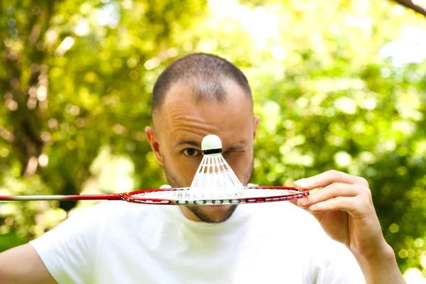 Genç adam göz kırpıyor ve yüzünü raket badmintonuyla kaplıyor. Güneşli bir günde park et. Yaşam tarzı, tatil, spor ve eğlence. Yakın plan..