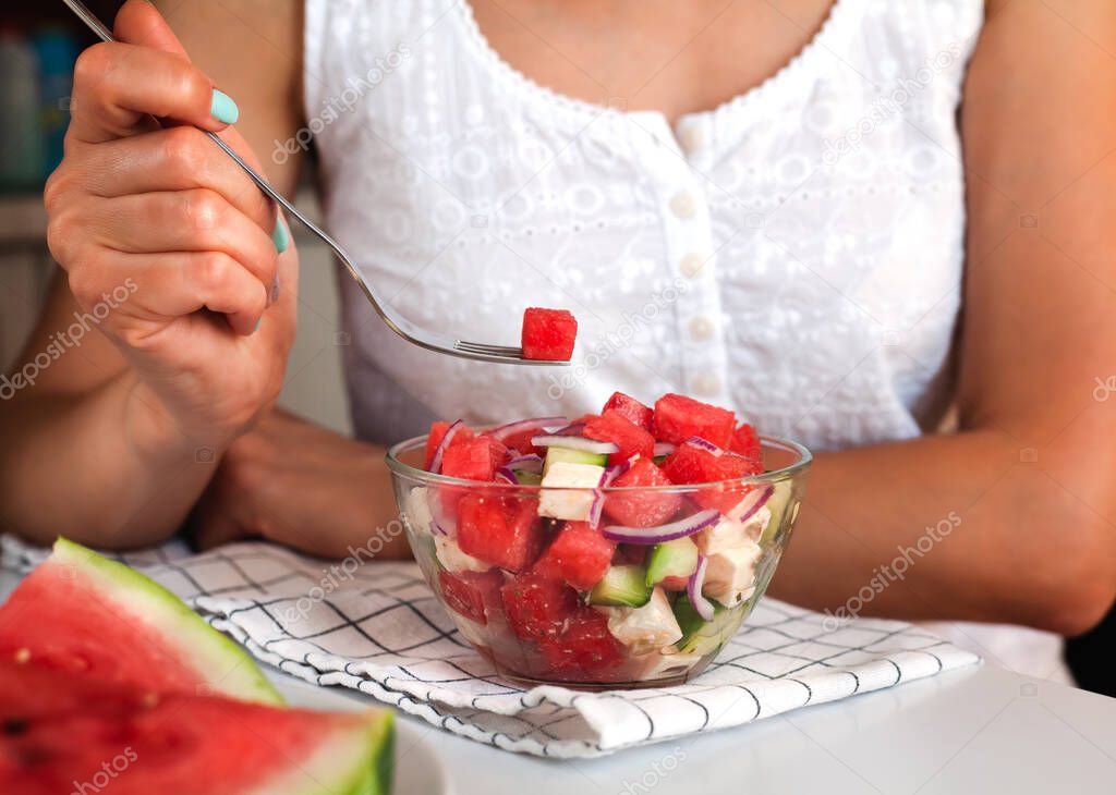 Young woman eating fresh juicy dietary salad with watermelon, feta cheese, cucumber and red onions on the kitchen at home. Close-up. Side view. Healthy food concept.