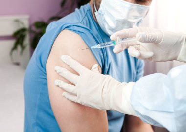 Covid-19 aşısı. Koruyucu lateks eldivenli ve koruyucu giysili bir doktor erkek hastaya iğne ya da aşı yapar. Sağlık hizmetleri. Yakın plan..