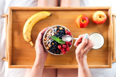 Kız yatağa oturur ve lezzetli ve sağlıklı bir kahvaltıda granola ve taze böğürtlen yer. Üst görünüm.