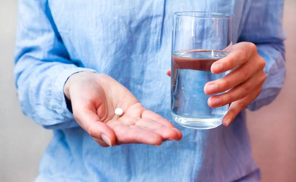 Woman in a blue blouse holds one pill and glass of water in hands. Taking painkiller medication or antibiotic, vitamins, antidepressant. Selective focus. Close-up.