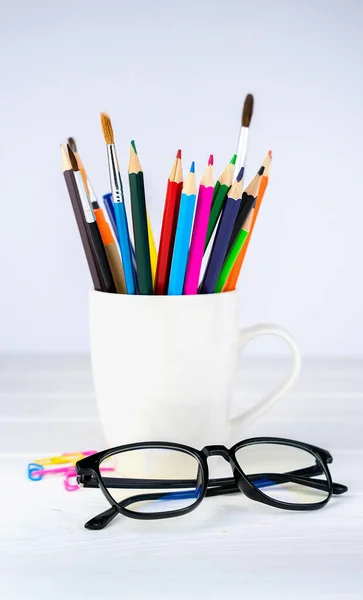 School supplies in a white mug and glasses on a white wooden background. Teacher\'s Day. Back to school. Close-up. Selective focus.