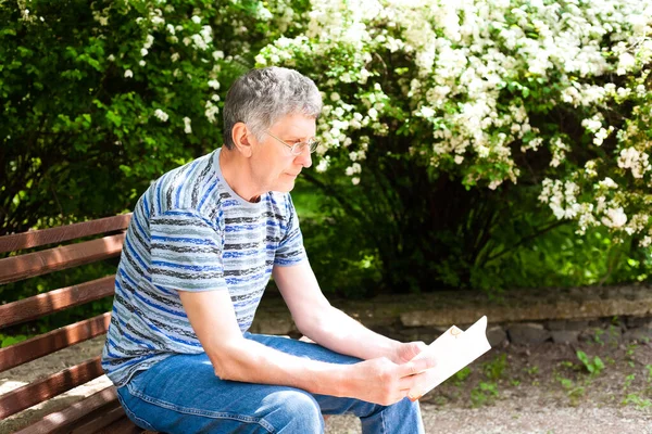 Senior man reading a book on a bench in the park in bright sunny day. Copy space.
