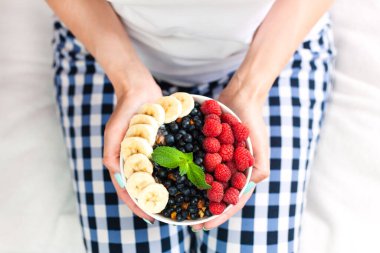 Female hands hold a bowl of granola with fresh berries and banana. Healthy breakfast. Close-up. Top view.