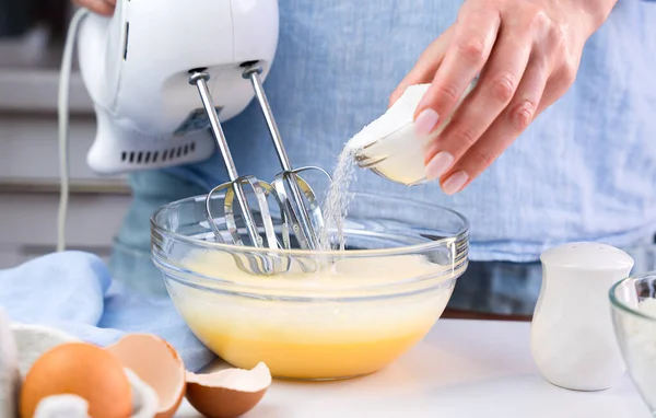 A woman beats eggs with a mixer and adds sugar while standing in the kitchen at home. Cooking and baking desserts at home. Close-up. Selective focus.