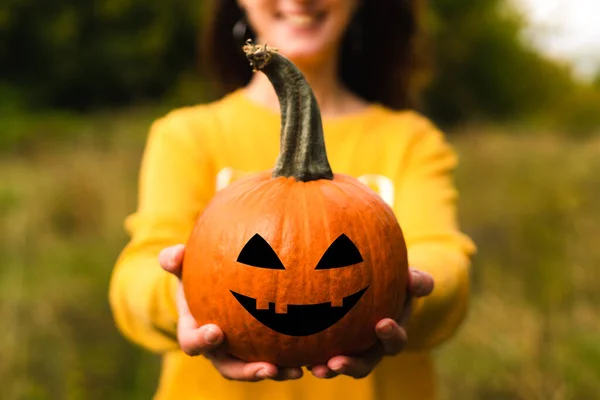 A smiling woman in a yellow sweatshirt holds an Halloween pumpkin with funny face. Halloween celebration concept. Close-up.