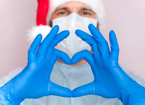 Doctor in a santa hat, medical mask and in protective gloves shows the symbol of the heart. Doctor care and love concept to patients. Love, care and safety symbol. Close-up.