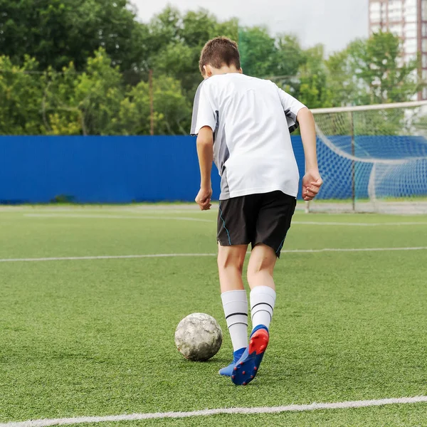 The boy loves to play football, trains with the ball on the artificial turf and scores a goal. The boy plays football.