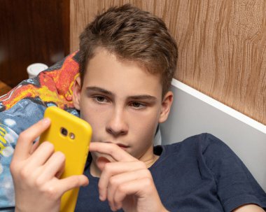 13-14 yaşlarında bir adam yatakta uzanıyor ve internet üzerinden arkadaşlarıyla iletişim kurarken gülüyor, cep telefonuyla oyun oynuyor..