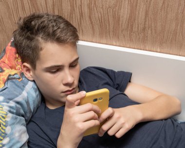 13-14 yaşlarında bir çocuk kanepede uzanıyor ve internette arkadaşlarıyla telefon aracılığıyla iletişim kurarken gülüyor, cep telefonuyla oyun oynuyor..