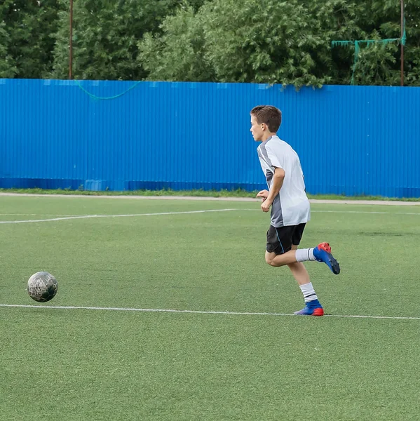 The boy loves to play football, trains with the ball on the artificial turf and scores a goal. The boy plays football.
