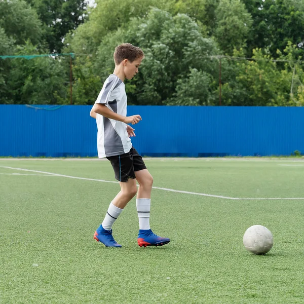 The boy loves to play football, trains with the ball on the artificial turf and scores a goal. The boy plays football.