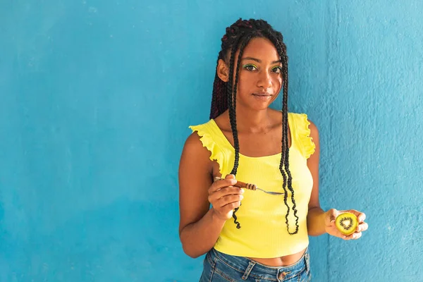 Siyah afro Latin Amerikalı sağlıklı kız örgülü ve kaşıkla kivi yiyor. Sarı gömlek ve kot etek. Arkadaki mavi duvar, turkuaz. Güzel, sıska kadın, yeşil sarı makyaj.