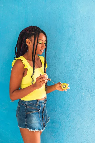 Siyah afro Latin Amerikalı sağlıklı kız örgülü ve kaşıkla kivi yiyor. Sarı gömlek ve kot etek. Arkadaki mavi duvar, turkuaz. Güzel, sıska kadın, yeşil sarı makyaj.