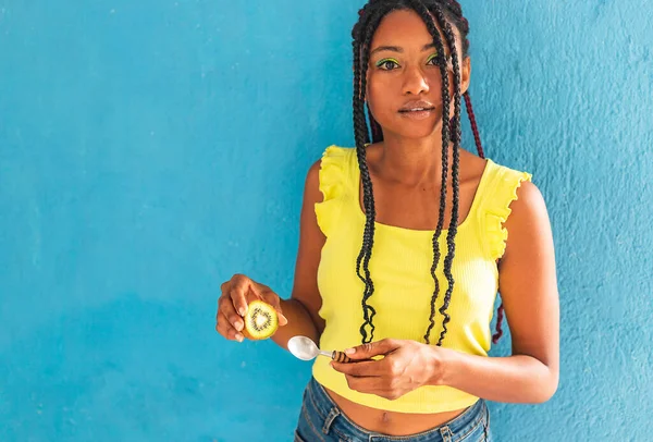 Siyah afro Latin Amerikalı sağlıklı kız örgülü ve kaşıkla kivi yiyor. Sarı gömlek ve kot etek. Arkadaki mavi duvar, turkuaz. Güzel, sıska kadın, yeşil sarı makyaj.