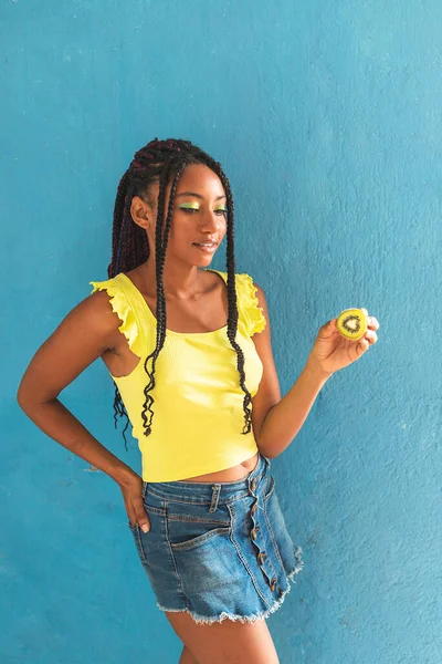 Siyah afro Latin Amerikalı sağlıklı kız örgülü ve kaşıkla kivi yiyor. Sarı gömlek ve kot etek. Arkadaki mavi duvar, turkuaz. Güzel, sıska kadın, yeşil sarı makyaj.