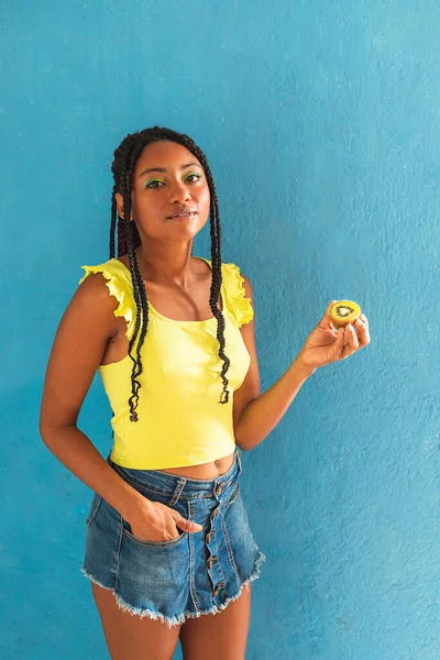 Siyah afro Latin Amerikalı sağlıklı kız örgülü ve kaşıkla kivi yiyor. Sarı gömlek ve kot etek. Arkadaki mavi duvar, turkuaz. Güzel, sıska kadın, yeşil sarı makyaj.