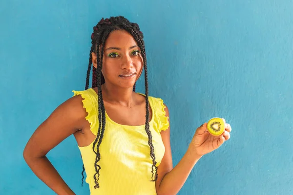 Siyah afro Latin Amerikalı sağlıklı kız örgülü ve kaşıkla kivi yiyor. Sarı gömlek ve kot etek. Arkadaki mavi duvar, turkuaz. Güzel, sıska kadın, yeşil sarı makyaj.
