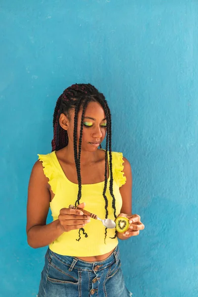 Siyah afro Latin Amerikalı sağlıklı kız örgülü ve kaşıkla kivi yiyor. Sarı gömlek ve kot etek. Arkadaki mavi duvar, turkuaz. Güzel, sıska kadın, yeşil sarı makyaj.