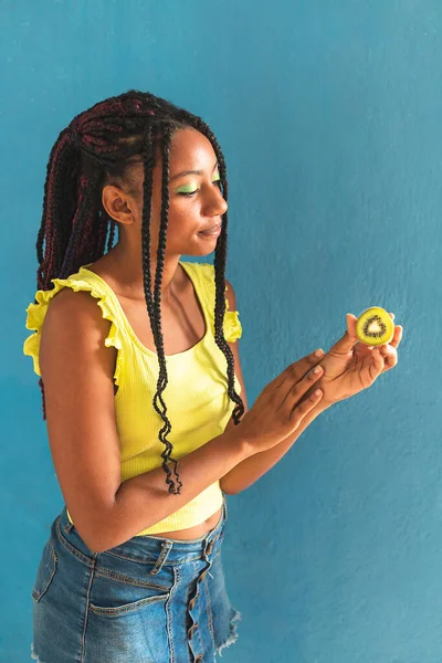 Siyah afro Latin Amerikalı sağlıklı kız örgülü ve kaşıkla kivi yiyor. Sarı gömlek ve kot etek. Arkadaki mavi duvar, turkuaz. Güzel, sıska kadın, yeşil sarı makyaj.