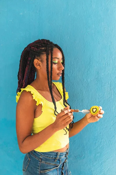Siyah afro Latin Amerikalı sağlıklı kız örgülü ve kaşıkla kivi yiyor. Sarı gömlek ve kot etek. Arkadaki mavi duvar, turkuaz. Güzel, sıska kadın, yeşil sarı makyaj.
