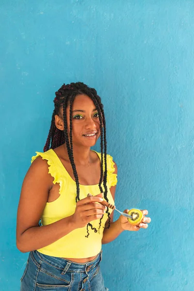 Siyah afro Latin Amerikalı sağlıklı kız örgülü ve kaşıkla kivi yiyor. Sarı gömlek ve kot etek. Arkadaki mavi duvar, turkuaz. Güzel, sıska kadın, yeşil sarı makyaj.