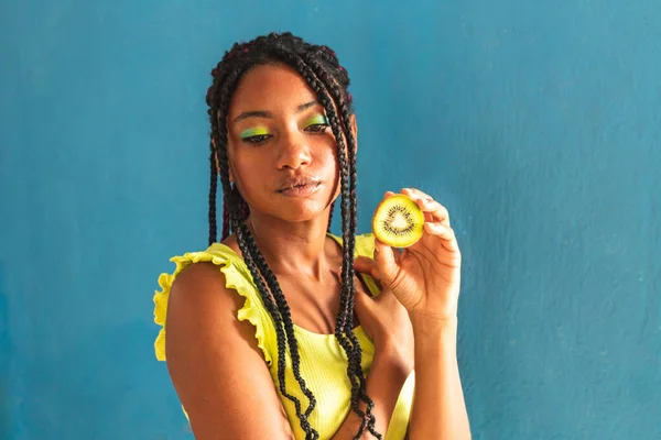 Siyah afro Latin Amerikalı sağlıklı kız örgülü ve kaşıkla kivi yiyor. Sarı gömlek ve kot etek. Arkadaki mavi duvar, turkuaz. Güzel, sıska kadın, yeşil sarı makyaj.