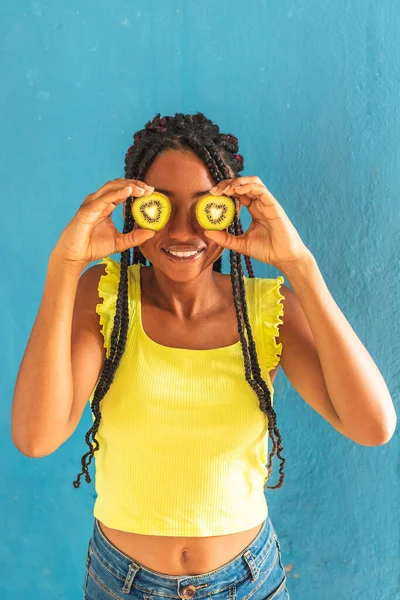Siyah afro Latin Amerikalı sağlıklı kız örgülü ve kaşıkla kivi yiyor. Sarı gömlek ve kot etek. Arkadaki mavi duvar, turkuaz. Güzel, sıska kadın, yeşil sarı makyaj.