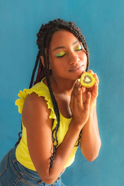 Siyah afro Latin Amerikalı sağlıklı kız örgülü ve kaşıkla kivi yiyor. Sarı gömlek ve kot etek. Arkadaki mavi duvar, turkuaz. Güzel, sıska kadın, yeşil sarı makyaj.