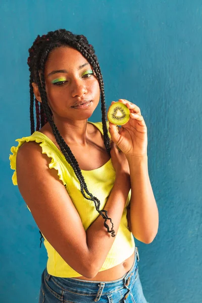 Siyah afro Latin Amerikalı sağlıklı kız örgülü ve kaşıkla kivi yiyor. Sarı gömlek ve kot etek. Arkadaki mavi duvar, turkuaz. Güzel, sıska kadın, yeşil sarı makyaj.