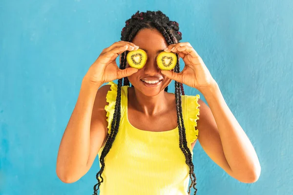 Siyah afro Latin Amerikalı sağlıklı kız örgülü ve kaşıkla kivi yiyor. Sarı gömlek ve kot etek. Arkadaki mavi duvar, turkuaz. Güzel, sıska kadın, yeşil sarı makyaj.
