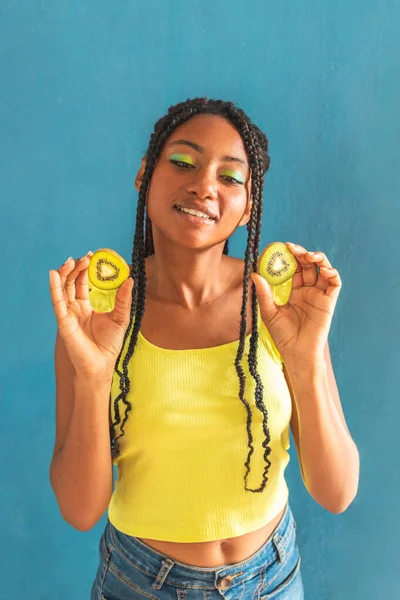 Siyah afro Latin Amerikalı sağlıklı kız örgülü ve kaşıkla kivi yiyor. Sarı gömlek ve kot etek. Arkadaki mavi duvar, turkuaz. Güzel, sıska kadın, yeşil sarı makyaj.