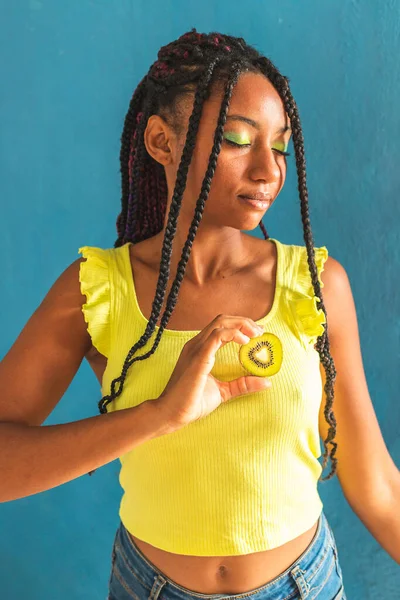 Siyah afro Latin Amerikalı sağlıklı kız örgülü ve kaşıkla kivi yiyor. Sarı gömlek ve kot etek. Arkadaki mavi duvar, turkuaz. Güzel, sıska kadın, yeşil sarı makyaj.