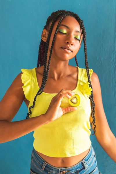 Siyah afro Latin Amerikalı sağlıklı kız örgülü ve kaşıkla kivi yiyor. Sarı gömlek ve kot etek. Arkadaki mavi duvar, turkuaz. Güzel, sıska kadın, yeşil sarı makyaj.