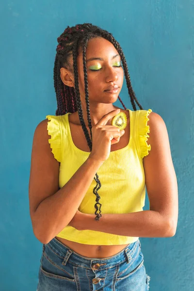Siyah afro Latin Amerikalı sağlıklı kız örgülü ve kaşıkla kivi yiyor. Sarı gömlek ve kot etek. Arkadaki mavi duvar, turkuaz. Güzel, sıska kadın, yeşil sarı makyaj.