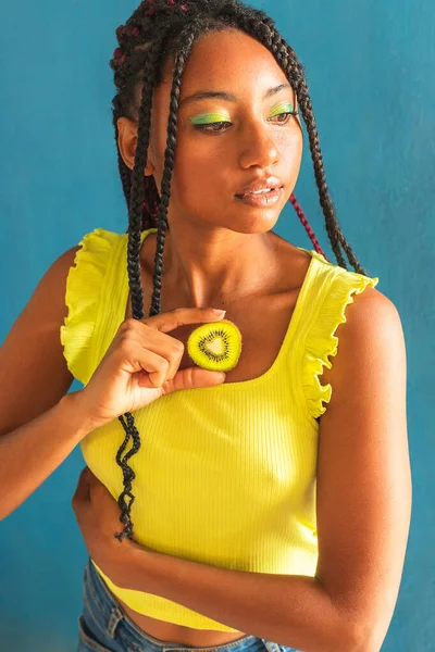Siyah afro Latin Amerikalı sağlıklı kız örgülü ve kaşıkla kivi yiyor. Sarı gömlek ve kot etek. Arkadaki mavi duvar, turkuaz. Güzel, sıska kadın, yeşil sarı makyaj.