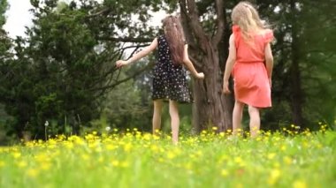 Unknown girls walk on green meadow at sunrise time. Rear view of female kid legs walking in city park. Unrecognizable child in pink dress playing in garden. High quality FullHD footage