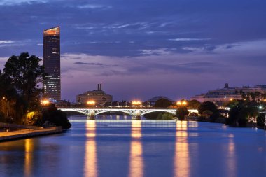 Gün batımında köprüden Sevilla 'daki Guadalquivir nehrinin manzarası, gökyüzünün mor ve mavi tonlarıyla ve nehirden yansıyan ışıklarla..