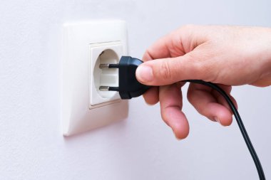 Plugging a black electrical plug with a womans hand into a white plastic socket on a wall. clipart
