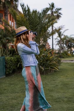 Şapkalı, uzun elbiseli ve kot ceketli çekici kadın muhabirin yan portresi palmiye ağaçları ve bitkilerle fotoğraf çekiyor..