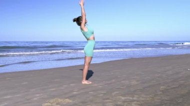 Yalnız bir kadının, sabahın erken saatlerinde, arka planda dalgalar olan ıssız bir plajda yoga yaparken çekilmiş yan görüntüsü. Sağlıklı yaşam ve vücut bakımı kavramı. 