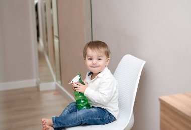 Beyaz bir sandalyede oturan küçük yakışıklı, beyaz bir sarışın çocuk, yeşil bir şişe suyla duygusal olarak oynuyor. Yumuşak odak.